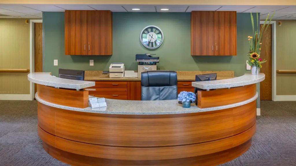 A modern reception desk with a semi-circular wooden design, featuring a black leather chair, computer monitors, a printer, and various office supplies. The green wall behind has a clock centered above the desk, with wooden cabinets and a flower arrangement on the right.