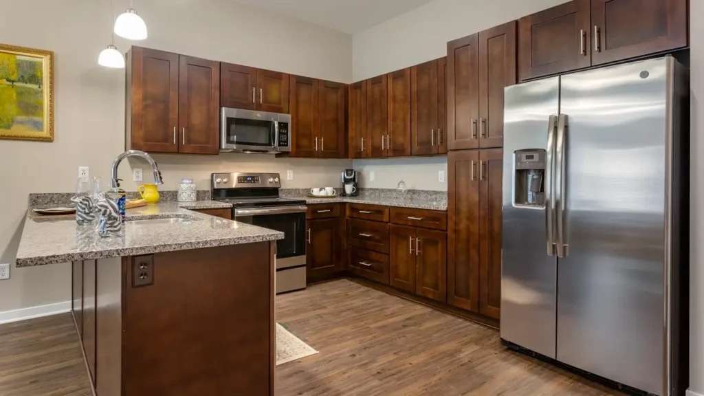 A modern kitchen featuring stainless steel appliances, including a refrigerator, oven, microwave, and dishwasher. The countertops are granite, and the cabinets are a rich brown wood. The kitchen has a wooden floor, and there is a painting on the left wall.