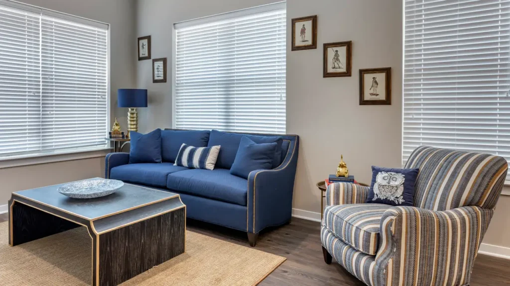 A cozy living room features a blue sofa adorned with patterned cushions, a striped armchair, and a dark coffee table with a decorative plate. The walls display framed pictures, and two windows with blinds provide natural light. A blue lamp sits beside the sofa.