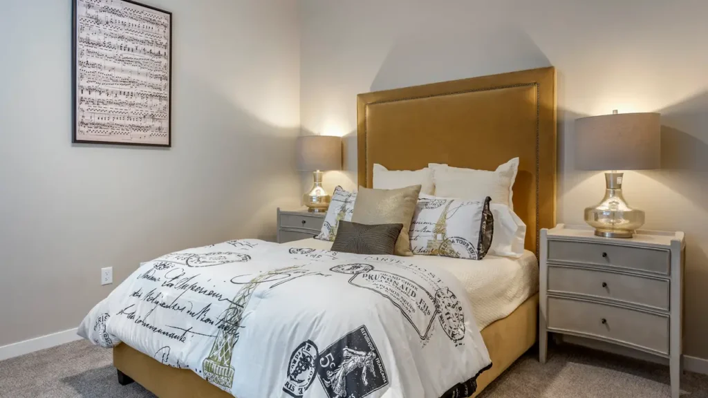 A cozy bedroom features a bed with a mustard-colored upholstered headboard, white bedding with black and gray script and stamp designs. Two gray nightstands, each with a silver lamp, flank the bed. A framed musical score hangs on the white wall to the left.
