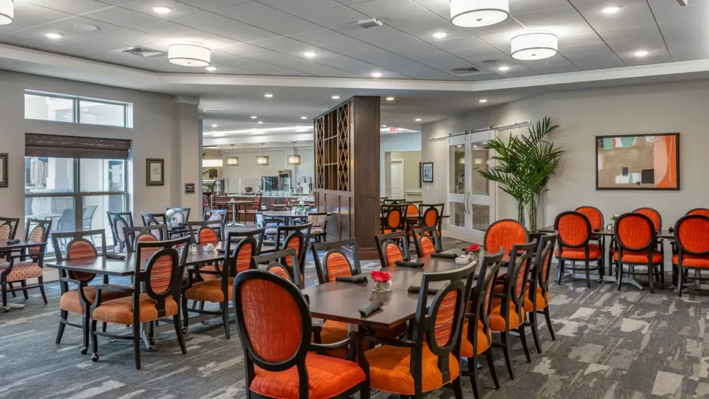 A spacious dining area with several tables and chairs featuring orange cushions. The room has modern lighting fixtures on the ceiling, large windows, and a central partition dividing the space. There are plants and framed artworks, creating a cozy ambiance.