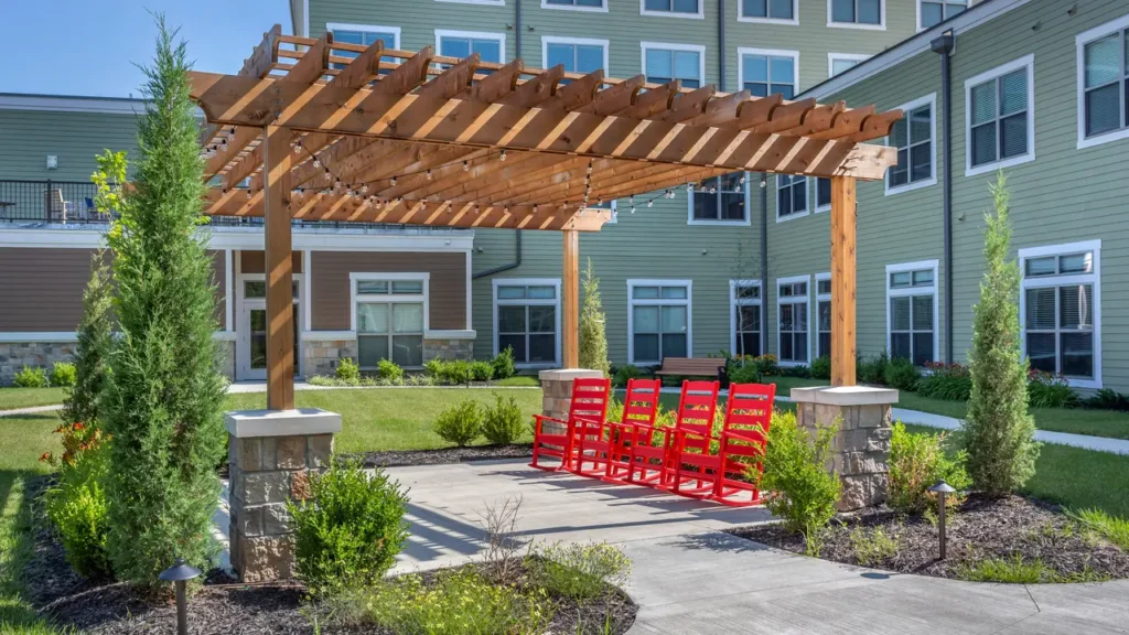 A well-maintained courtyard with a wooden pergola, adorned with string lights. Underneath, there are four bright red rocking chairs on a concrete platform, surrounded by greenery. In the background are multi-story buildings with large windows and beige and green exteriors.