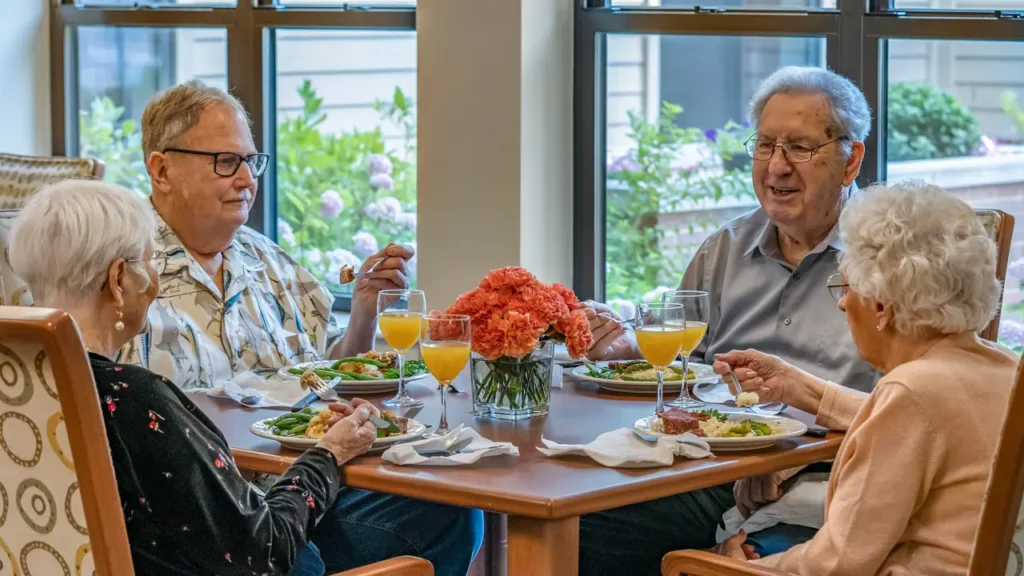 Four elderly people are dining together at a table set with meals and beverages. The table is decorated with a bouquet of orange flowers. They are seated in a brightly lit room with large windows showing greenery outside, engaging in conversation and enjoying their meal.