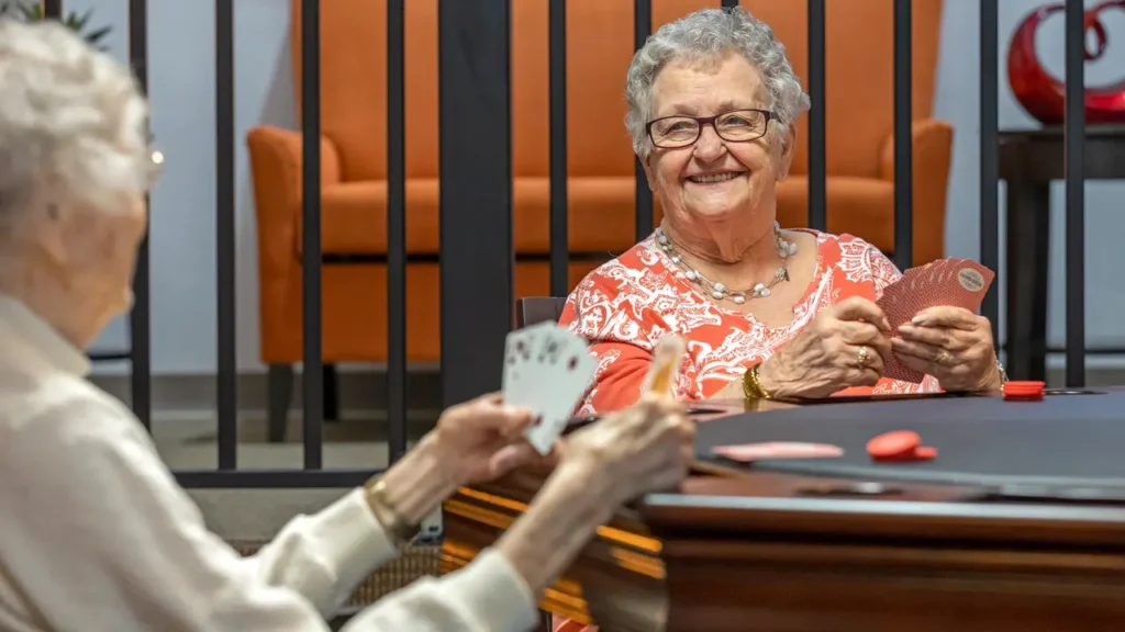 Two elderly women are sitting at a table playing cards. The woman on the right, wearing glasses and a patterned top, smiles while holding her cards. The woman on the left, also holding cards, is partially visible. An orange chair and a black railing are in the background.