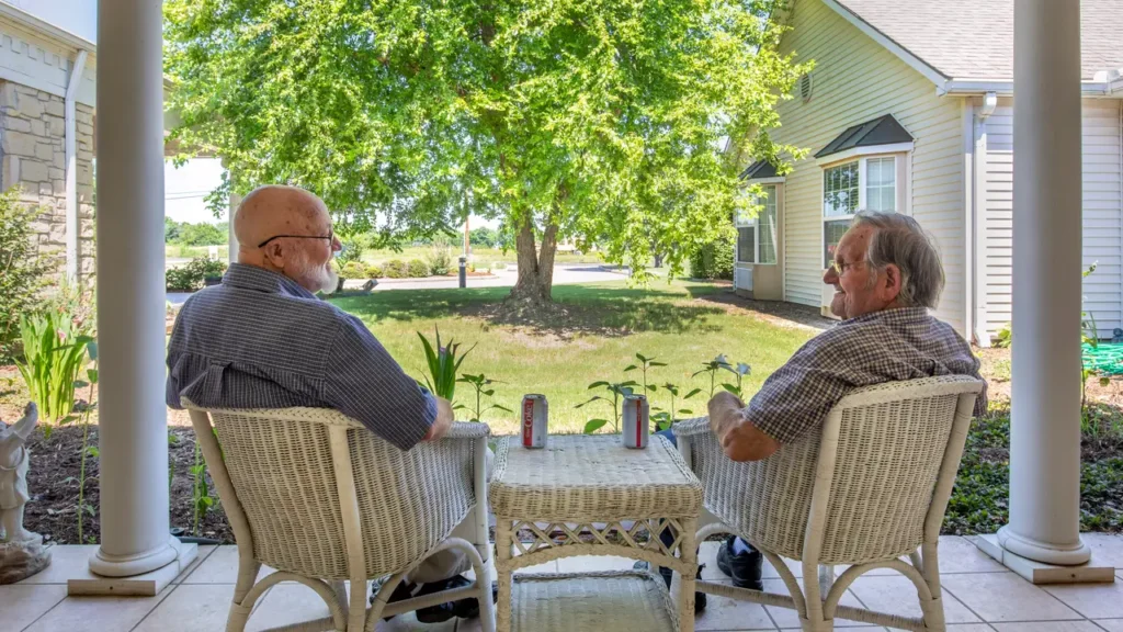 Two older men sit and chat on white wicker chairs on a porch, with a wicker table holding two cans in between them. They are surrounded by a lush green lawn and trees, with a light yellow house visible in the background.