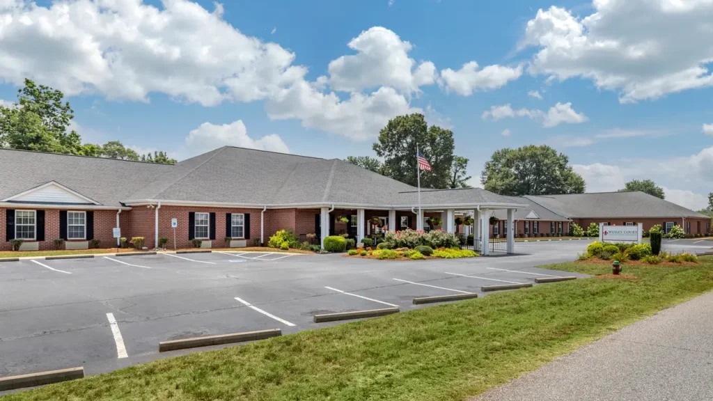 A brick building with a gray roof sits under a blue sky with scattered clouds. An American flag is on a pole near the entrance, which features a white portico. The surrounding area is landscaped with bushes and trees, and an empty paved parking lot is in the foreground.