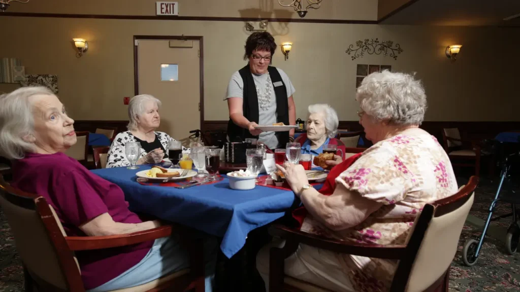 A group of four elderly women sit around a dining table with blue tablecloths, enjoying a meal together. A staff member stands beside the table, serving food from a tray. The room is well-lit, and the atmosphere is warm and friendly.