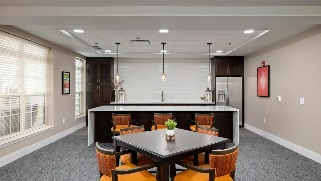 A modern kitchen and dining area featuring a dark wood island with four matching chairs, a square dark wood table with orange-cushioned chairs, pendant lights, stainless steel refrigerator, white backsplash, and neutral-colored walls. A small plant adorns the table.