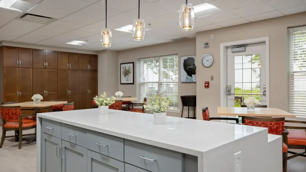 A modern kitchen with a large white island countertop. Three pendant lights hang above the island, which has two plants on it. In the background, there is seating with red chairs, wooden cabinets, and a clock on the wall. Sunlight comes through the window.