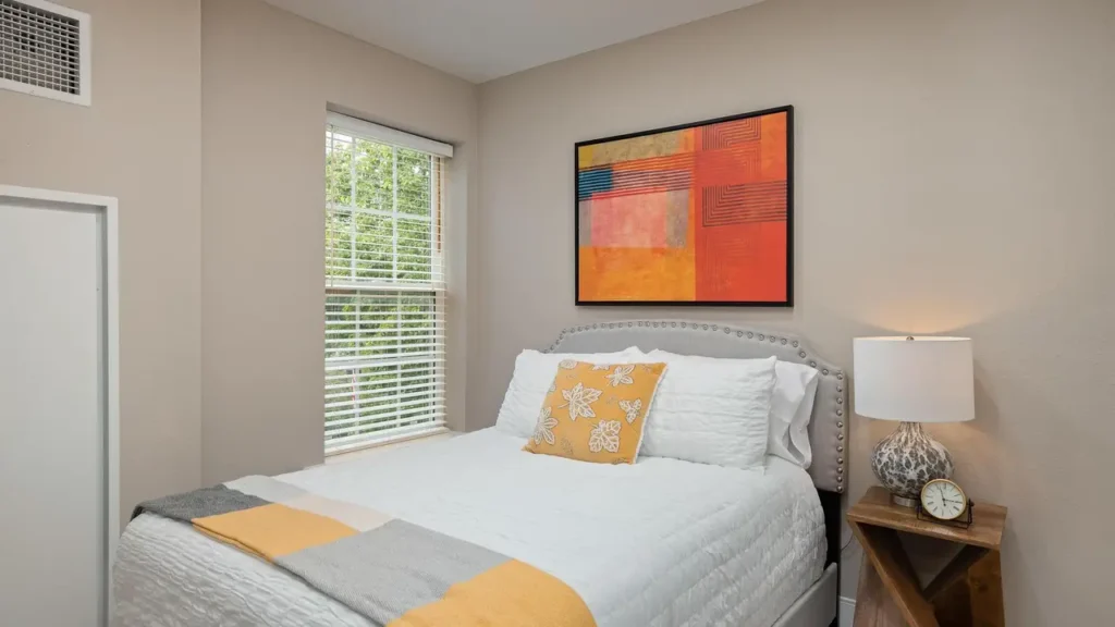 A cozy bedroom with beige walls features a bed adorned with white and yellow bedding. Above the bed hangs a colorful abstract painting with shades of red, orange, and blue. A wooden nightstand with a lamp and decorative vase sits beside the bed near a window.