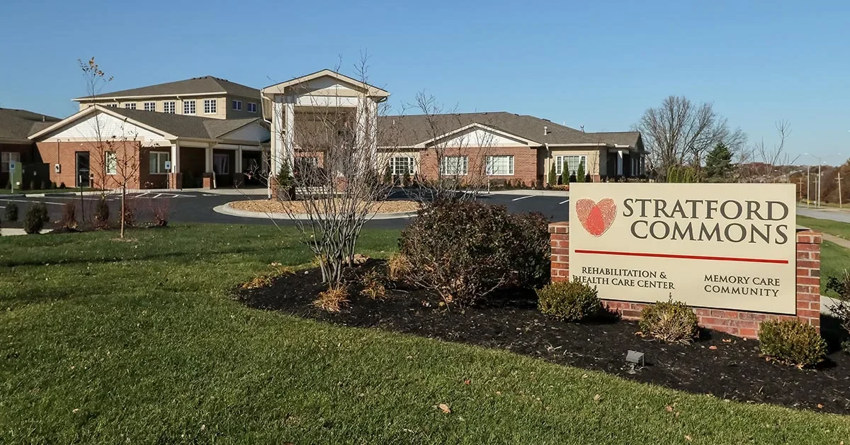 A modern brick building with white accents houses the Stratford Commons Rehabilitation & Health Care Center, a memory care community. The facility's sign is prominently displayed in the foreground, surrounded by neatly maintained lawns and bare trees.