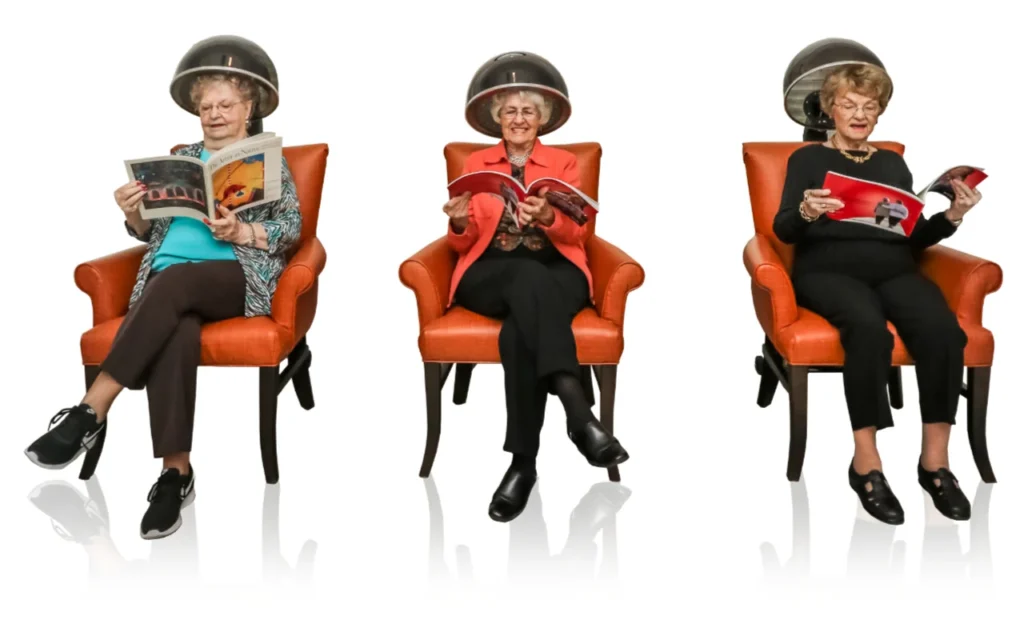 Three elderly women are enjoying their senior living by sitting under old-fashioned hair dryers, reading magazines. They each sit in an orange chair against a white background. The woman on the left wears a blue outfit, the middle woman a red one, and the woman on the right dresses in black.