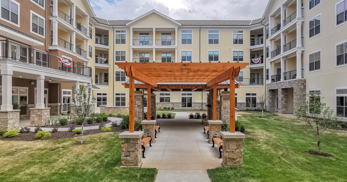 A courtyard of a senior living apartment complex with beige four-story buildings surrounding it. The courtyard features a wooden pergola with benches underneath, neatly landscaped garden beds, and a well-maintained lawn. Some balconies display potted plants and decorations.