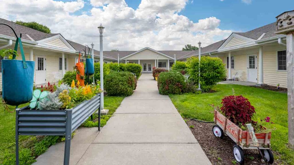 A concrete pathway runs through a landscaped courtyard between light-colored, single-story buildings with white trim. Flower beds and planters, including a red wagon with plants, decorate the area under a partly cloudy sky.