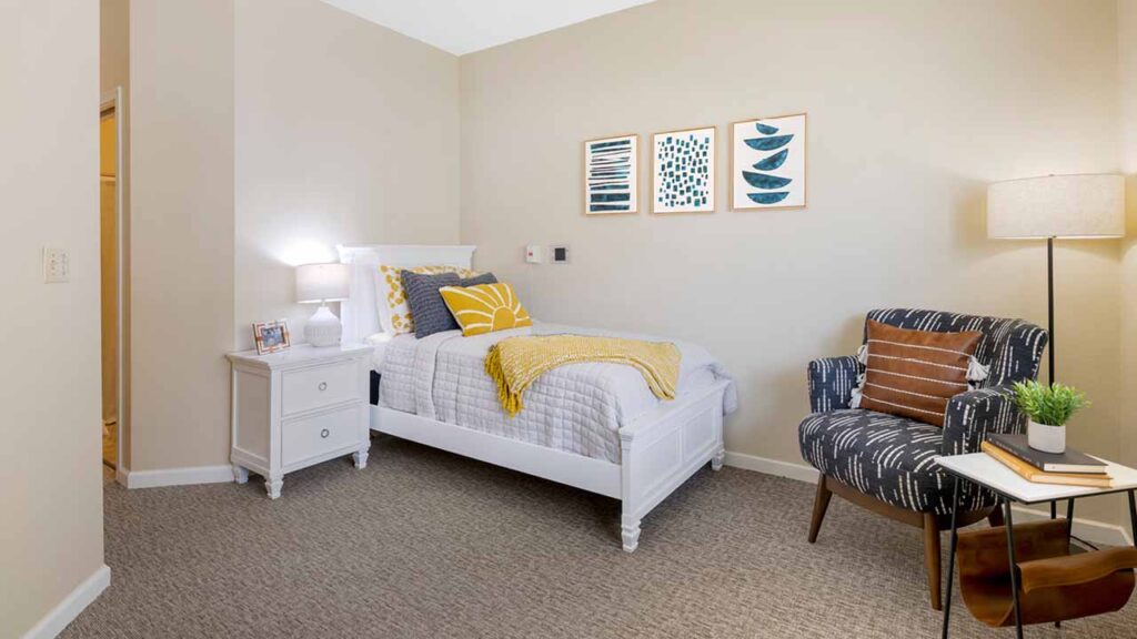 A cozy bedroom with a white single bed, yellow and gray bedding, a white nightstand with a lamp, wall art above the bed, an armchair with patterned upholstery, a floor lamp, and a small round side table with a plant.