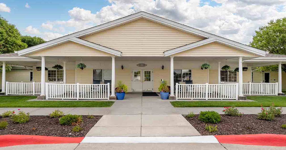 Single-story beige building with a wide front porch, white railings, hanging plants, and blue flower pots by the entrance, set against a partly cloudy sky, with landscaped walkway and trimmed bushes in front.