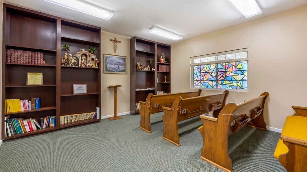 Small chapel room with wooden pews, built-in bookshelves, religious statues, a crucifix, a podium, framed art, and a window with colorful stained glass patterns. The room is well-lit with fluorescent lights.