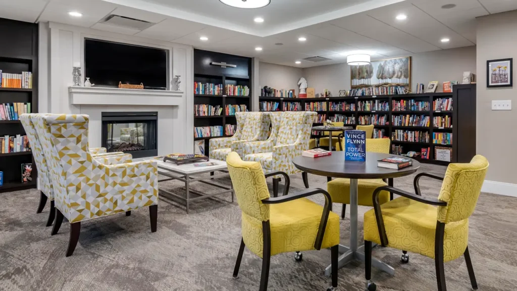 A cozy library room with bookshelves lining the walls, filled with books. The space features a fireplace with a TV above it, surrounded by armchairs and a large coffee table. A small round table with chairs and books is in the foreground, under bright ceiling lights.