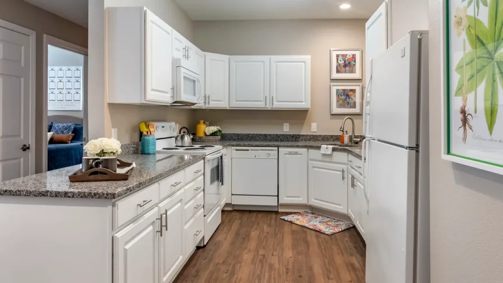 Modern kitchen with white cabinets and appliances, granite countertops, and wood laminate flooring. Decorative art pieces and a framed plant print adorn the walls. A tray with flowers and a colorful rug add a touch of color to the space.