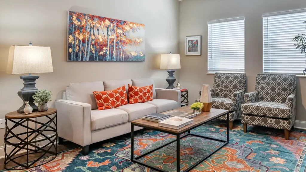 A cozy living room featuring a light grey sofa adorned with two orange patterned cushions. Above the sofa hangs a colorful abstract painting of trees. The room also has a patterned armchair, two lamps, a wooden coffee table, and a vibrant multicolored rug. Natural light fills the space through the window blinds.