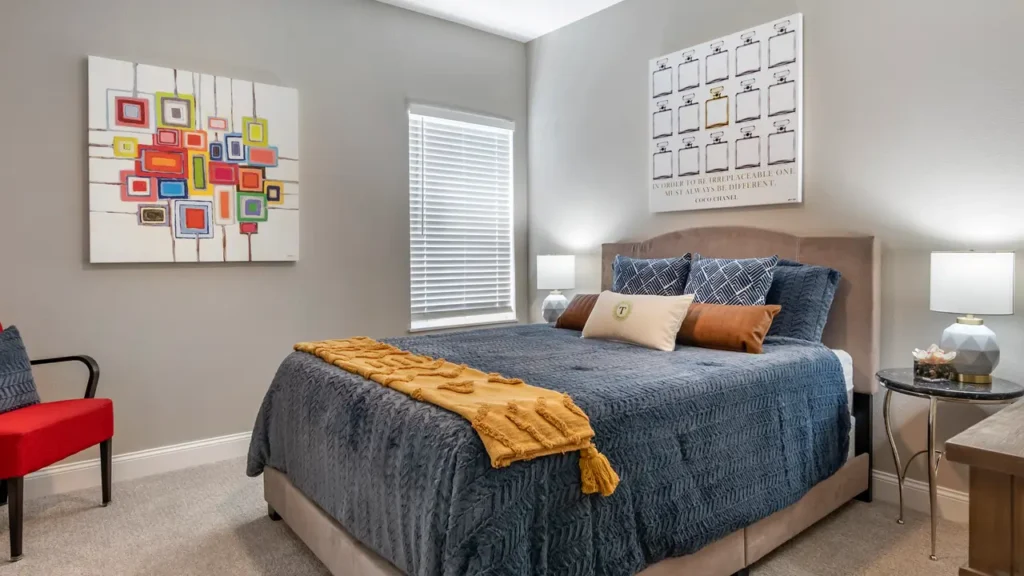 A bedroom features a neatly made bed with a dark blue quilt, several decorative pillows, and a mustard-colored throw blanket. The walls are adorned with colorful abstract art and a framed print. A window with white blinds lets in natural light.