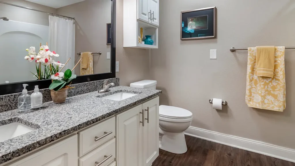 A modern bathroom with white cabinets, a granite countertop, and a single sink. Above the sink is a large mirror. To the right are a toilet, a towel ring with a yellow towel, and a wall-mounted artwork. The floor is made of dark wood.