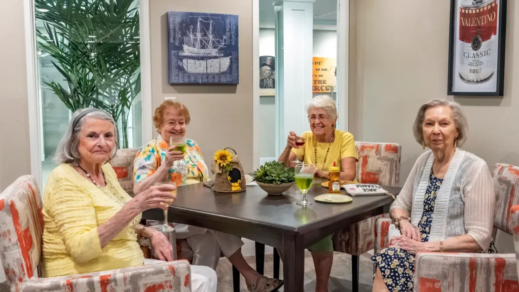 Four elderly women sit around a table, smiling and raising glasses in a toast. They are in a bright, cozy room with cheerful decor, including plants and nautical-themed artwork. Two of them wear yellow tops, the others are dressed in light-colored clothing.