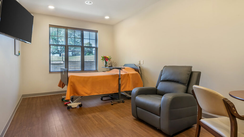A hospital or senior care room with a single bed covered in an orange blanket, a bouquet of flowers on a table, a reclining chair, and a window with blinds letting in daylight.
