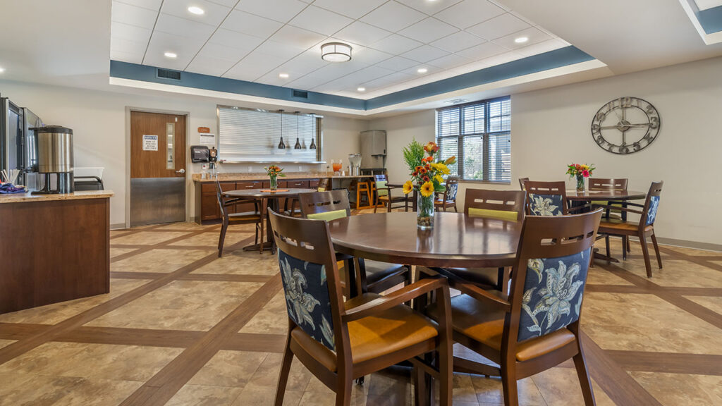 A well-lit dining area with wooden tables and chairs, floral centerpieces, a large wall clock, beverage station, and large windows letting in natural light. The space has a clean, modern design.