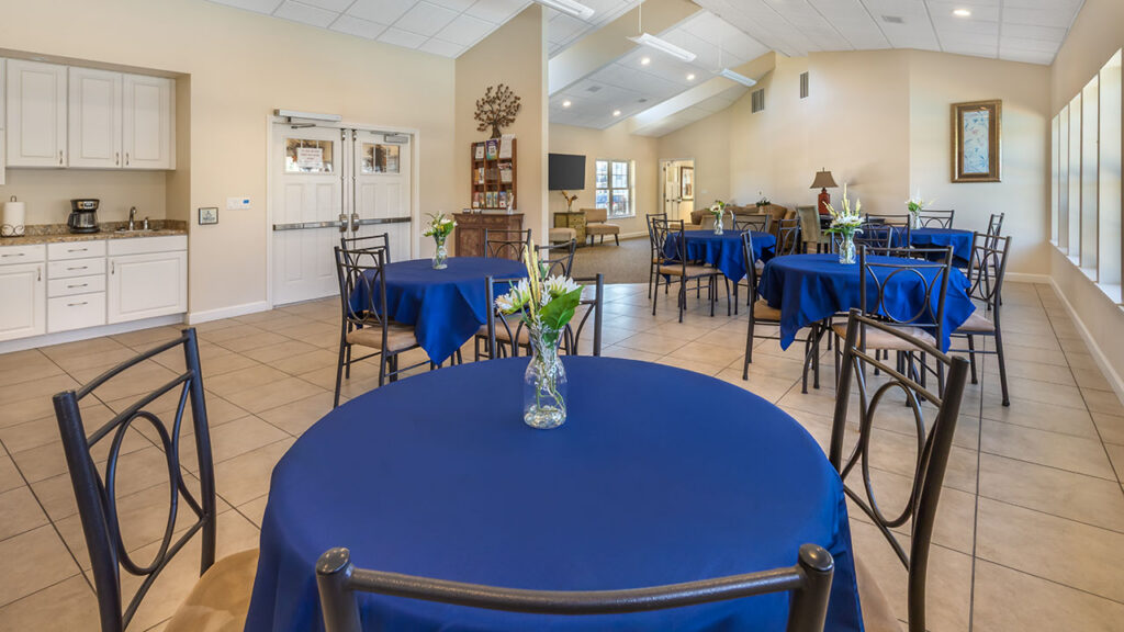 A bright, spacious room with round tables covered in blue tablecloths, each with a small vase of flowers. The room has tiled floors, white cabinets, and large windows letting in natural light.