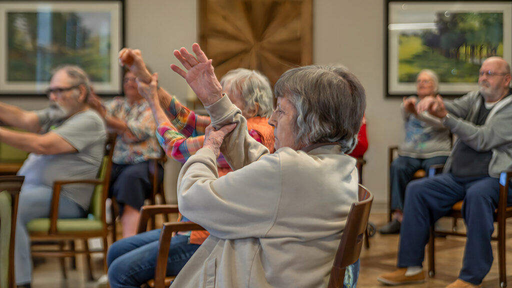 A group of elderly people sit in chairs indoors, participating in a seated exercise class with arms raised. The background shows framed artwork and wooden wall decor.