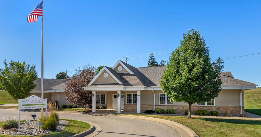 Single-story beige building with a gabled roof and white trim, surrounded by trees and landscaping. An American flag flies on a tall pole near a sign that reads “Grand Meadows.” The sky is clear and blue.