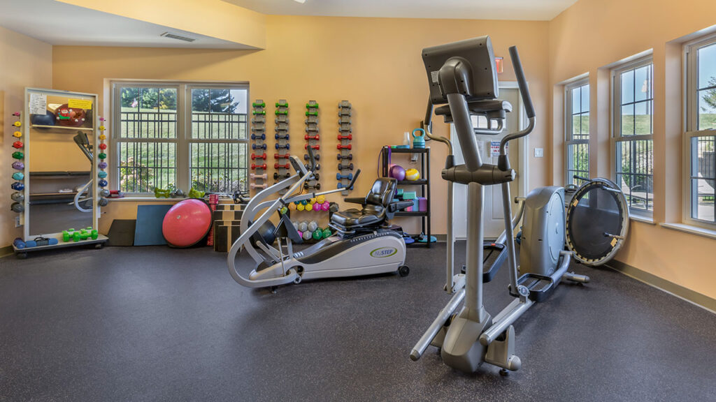 A small gym room with cardio machines, dumbbells, exercise balls, mats, and resistance bands, set against yellow walls and large windows letting in natural light.