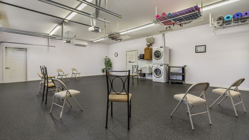 A spacious, brightly lit room with several chairs arranged in a distanced circle. There are shelves with yoga mats, a washer and dryer, a potted plant, and minimal décor on the light-colored walls.