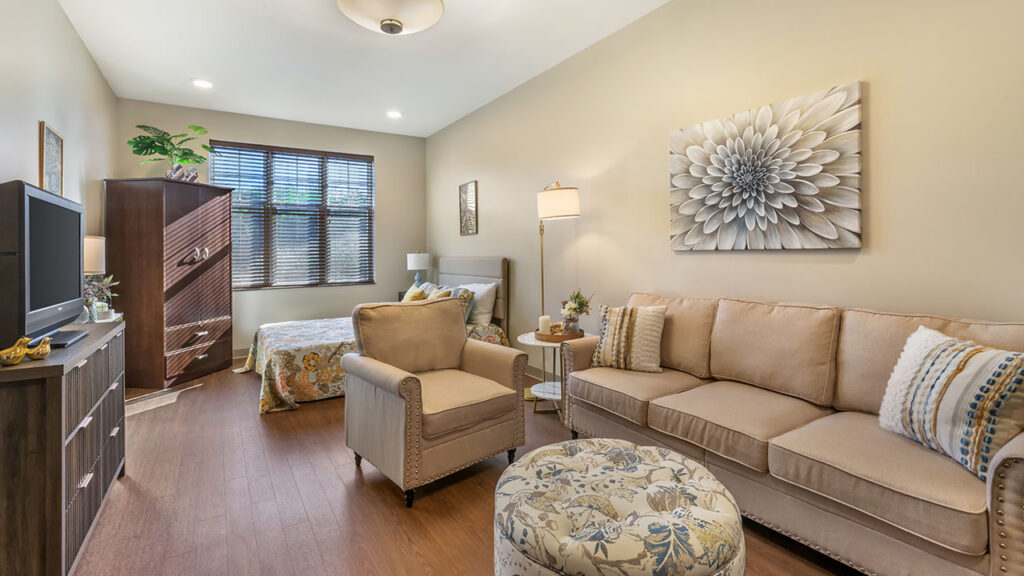 A cozy studio apartment with a bed, beige sofa, armchair, patterned ottoman, TV on a stand, wooden wardrobe, and a large flower artwork on the wall. The room has warm lighting and a window with blinds.