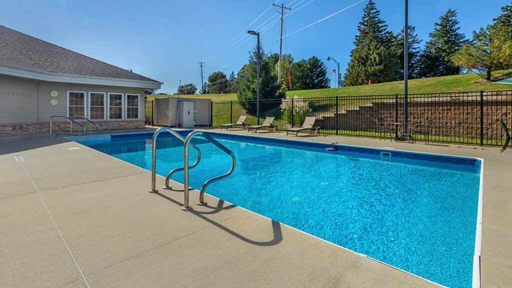 Outdoor swimming pool with clear blue water, surrounded by a beige concrete deck. There are metal handrails, several lounge chairs, a building to the left, and a grassy area with trees and a fence in the background.