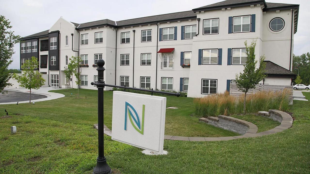 A modern three-story apartment building, designed for assisted living and memory care, boasts a landscaped lawn with trees. A sign with a stylized letter "N" sits on a black post out front. The structure features a blend of white and dark panels, some windows adorned with awnings.