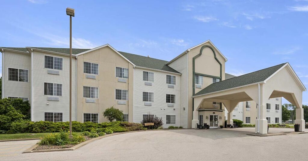 A three-story beige and white hotel building with multiple windows, a covered entrance, green roof, and surrounding landscaping on a sunny day.