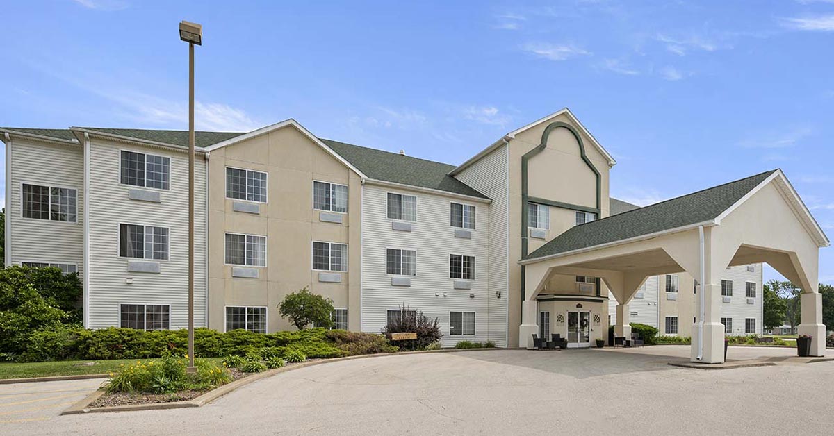 A three-story beige and white hotel building with multiple windows, a covered entrance, green roof, and surrounding landscaping on a sunny day.
