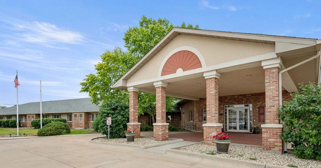 Single-story brick building with a covered entrance supported by columns, surrounded by landscaped bushes and trees. An American flag is on a pole to the left, and the sky is blue with scattered clouds.