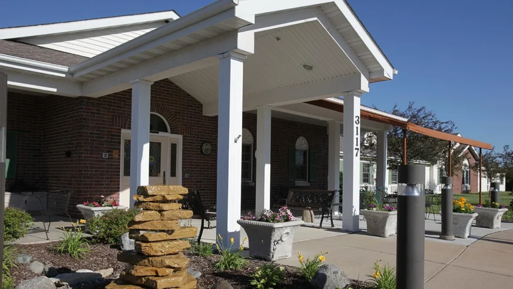 A brick building with a white gabled entrance featuring four columns. A stack of stones serves as a focal point in a landscaped garden area in front, which includes various plants and flowers. The assisted living and memory care facility is marked with the number 