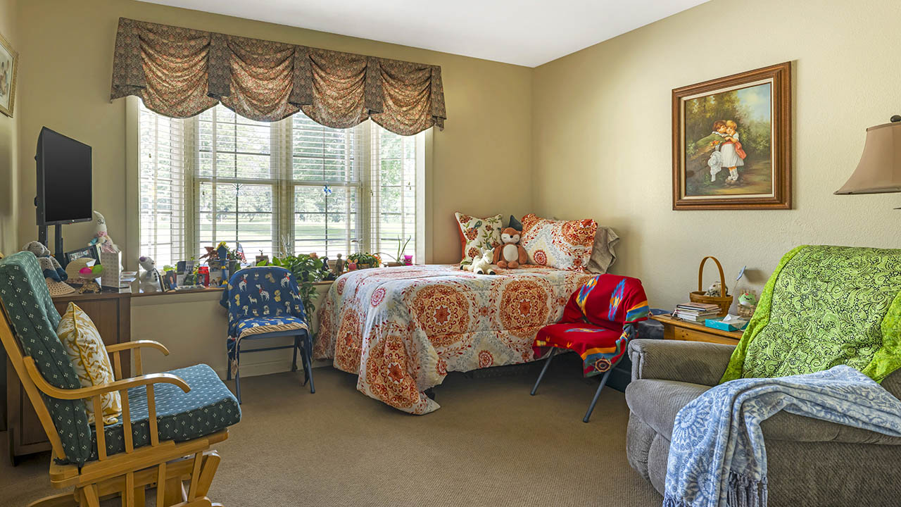 A cozy bedroom with a bed by a large window, colorful bedding, plush toys, a rocking chair, an armchair with throws, a side table with a lamp, framed artwork on the wall, and various decorative items.
