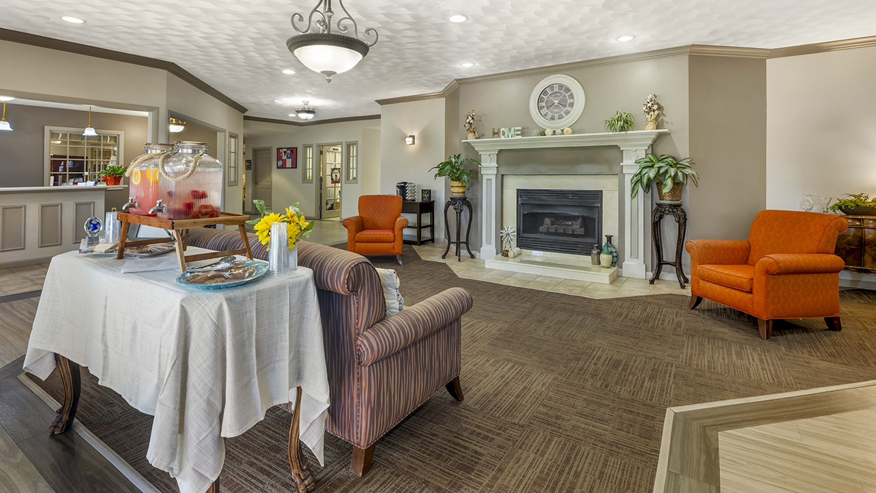 A cozy, well-lit living room with a fireplace, orange armchairs, and a table with refreshments. Plants and decorative items adorn the mantel, and a large ceiling light hangs above.