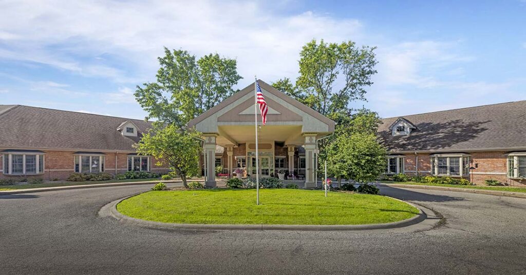 A brick building with a covered entrance and an American flag on a grassy island in front, surrounded by trees and a circular driveway under a blue sky.