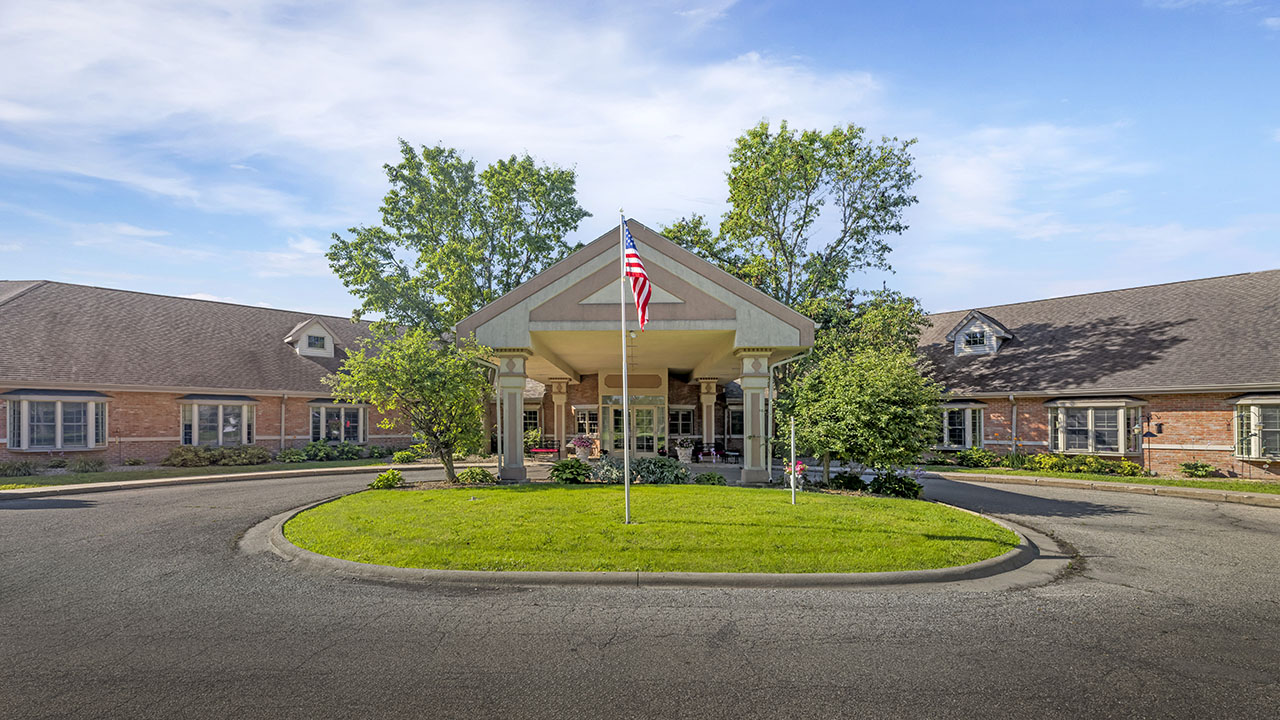 A brick building with a covered entrance and circular driveway, featuring an American flag on a pole in the grassy center, surrounded by trees and bushes under a clear blue sky.