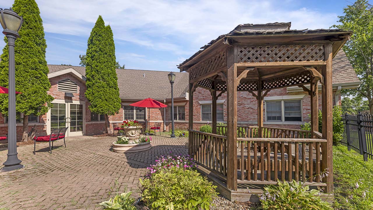 A wooden gazebo stands in a brick patio surrounded by flowers and greenery, with a red brick building, red umbrellas, and outdoor seating in the background on a sunny day.