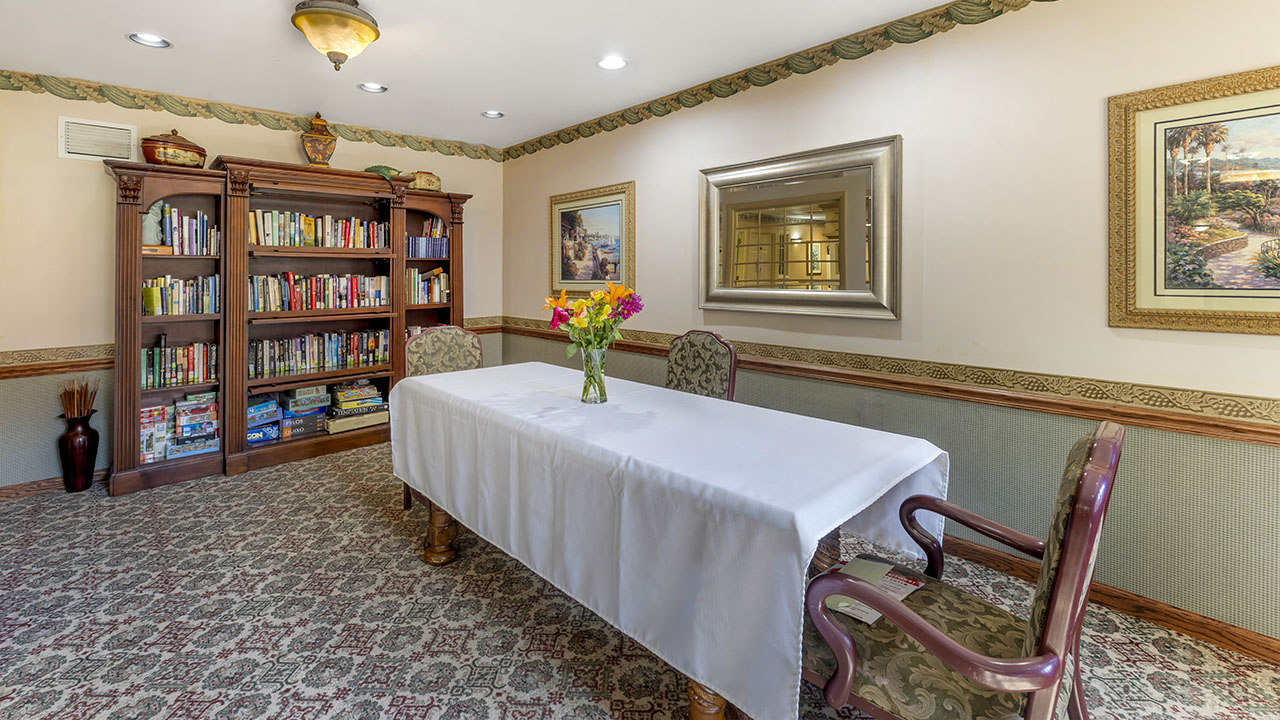 A cozy room with a bookshelf filled with books and board games, a table covered with a white tablecloth, three ornate chairs, wall art, and a vase of colorful flowers on the table.