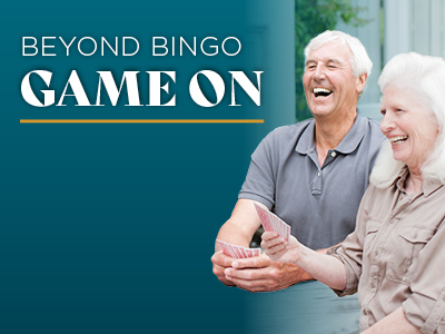Two older adults laugh and play cards together at a table, enjoying the vibrant Senior Living community. The text on the left reads, 