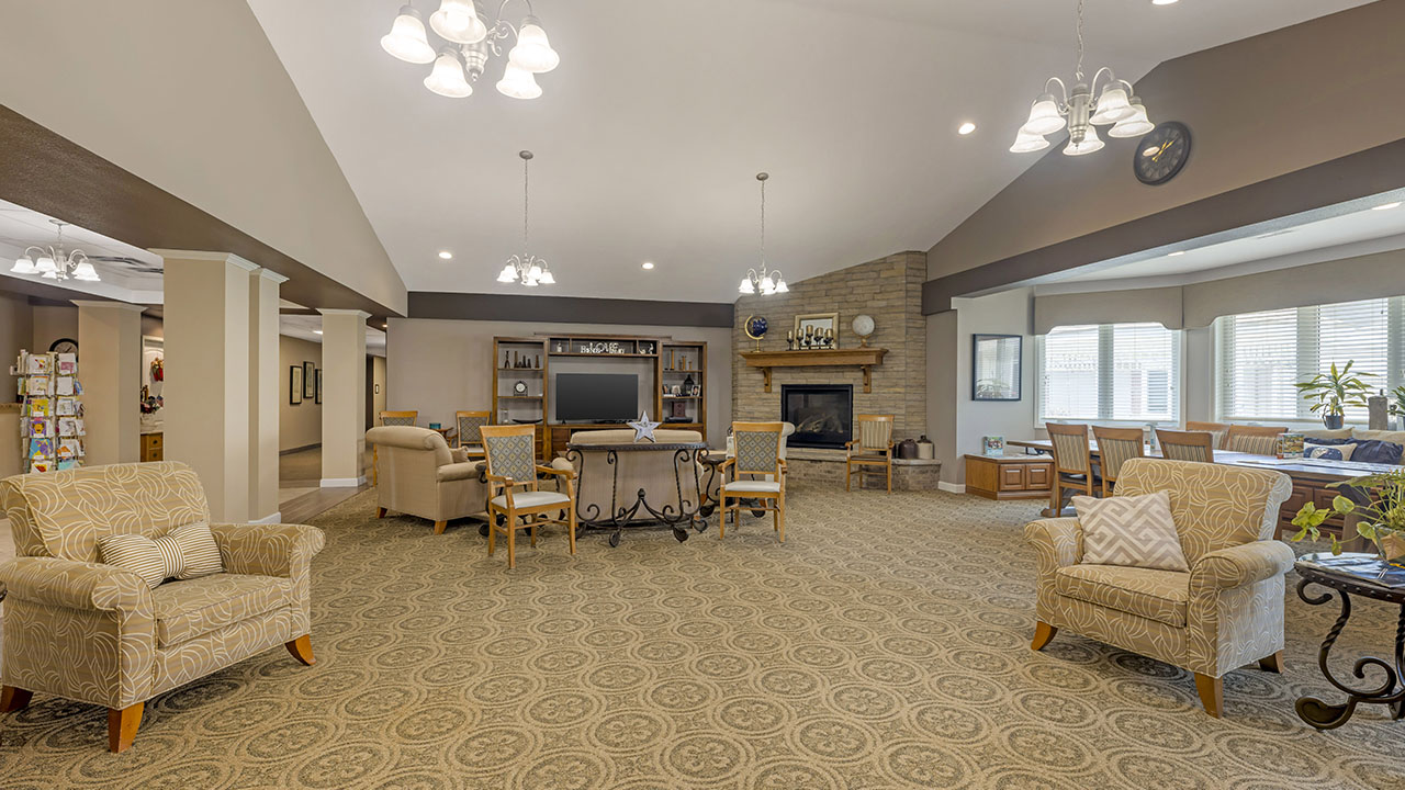 Spacious living room with patterned carpet, armchairs, sofas, a large TV, fireplace, bookshelves, dining area, and windows letting in natural light. Warm, neutral tones and modern lighting fixtures throughout.