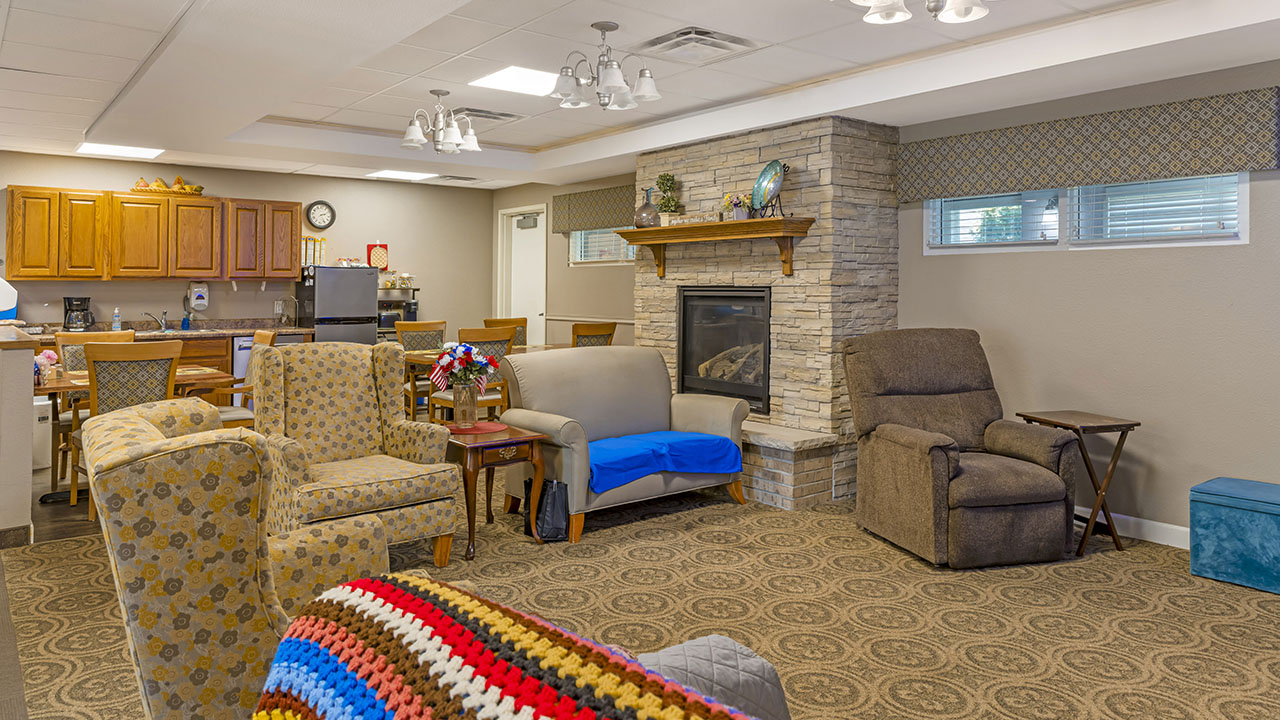 A cozy living room with patterned armchairs, a recliner, a fireplace with a stone surround, and a kitchen area in the background. The room has warm lighting, a carpeted floor, and decorations like flowers and a crocheted blanket.